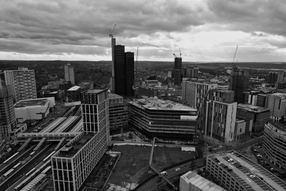 A black and white aerial photograph of a city skyline showing tall, modern office buildings and skyscrapers, some of which are under construction with cranes visible on top. The scene includes multiple mid-rise structures with glass and concrete facades, along with a transportation infrastructure featuring train tracks and platforms in the foreground. The sky is filled with dense clouds, casting a subdued light over the urban environment. The view captures the dense cityscape extending into the distance, highlighting ongoing development and construction activity, typical of a busy metropolitan area. This image reflects urban growth relevant to the logistics and planning involved in house removals or furniture transport services provided by Croydon Man and Van, illustrating the kind of environment where home relocations and moving logistics take place.