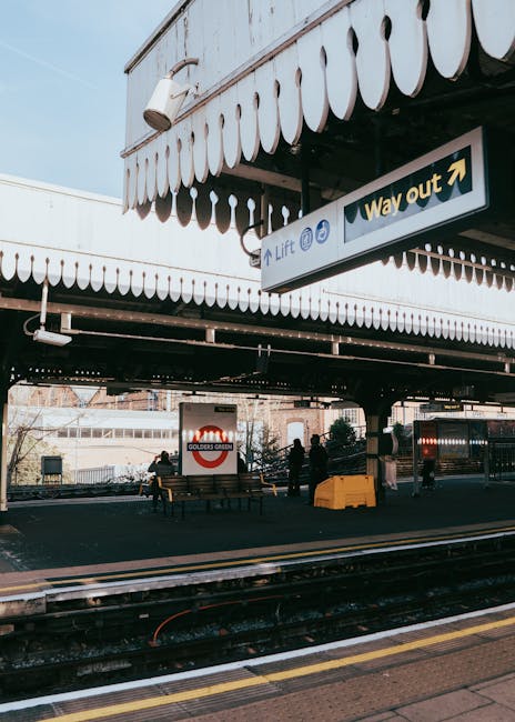 The image shows the exterior of East Croydon station, with a covered pedestrian area featuring a decorative white scalloped awning. Underneath, there is a paved space with two wooden benches, one occupied by a person, and several groups of people waiting either on the platform or near the station entrance. A prominent sign indicates the station's name 'GOLDERS GREEN' in red and white, and there is a 'Way out' sign with an arrow pointing to the right, guiding passengers towards the station exit. The railway tracks run parallel to the platform, and in the foreground, a yellow protective barrier is positioned on the pavement. The scene appears during daylight hours, with natural lighting illuminating the area. This setting relates to the station's infrastructure, which may be relevant for users planning home relocation or furniture transport via local train services, supported by companies like Croydon Man and Van for moving and packing logistics.