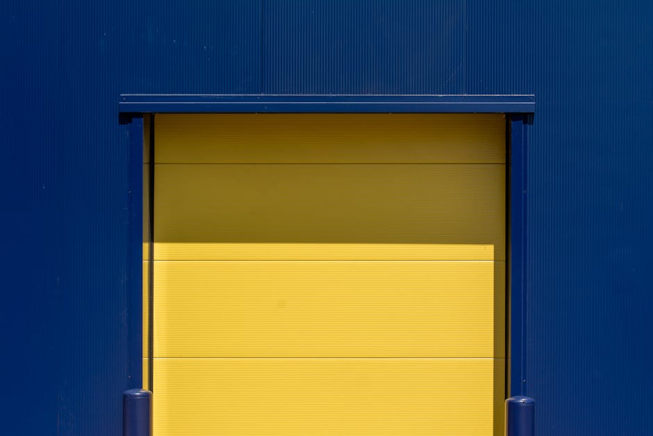 Close-up of a yellow loading bay door set within a blue industrial building exterior, with the door partially open revealing a glimpse of the interior. The door appears to be made of horizontally ribbed metal and is situated on a pavement area. At the bottom, small blue bollards are visible, likely to protect the entrance. The surrounding blue metal cladding features vertical ridges, indicating a warehouse or commercial property used for home relocation and furniture transport services. This scene captures the tight loading bay access commonly encountered during residential or commercial removals, highlighting the importance of precise packing and loading techniques employed by Croydon Man and Van for efficient moving operations.