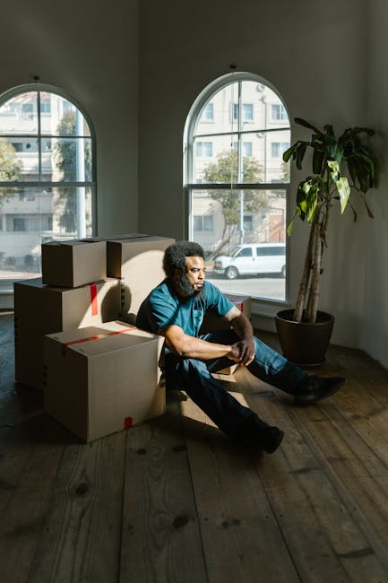 A man with curly dark hair and a beard, wearing a blue t-shirt and dark jeans, is sitting on a wooden floor inside a bright room with large arched windows that let in natural light. He is surrounded by several cardboard boxes, some sealed with red tape, indicating packing for a home relocation or moving process. The boxes vary in size and are stacked or placed nearby, suggesting they are ready for transport or unloading. There is a tall potted plant with broad green leaves positioned next to the window, adding a decorative element to the scene. The room appears to be in a residential property, with visible street view outside the windows, showing parked cars and a building across the street. The lighting highlights the interior space and the man's contemplative expression, reflecting a moment during the packing or loading phase of furniture transport, as part of house removal services provided by Croydon Man and Van.