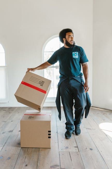 A man with dark, curly hair and a beard is inside a bright, spacious room with light-colored walls and wooden flooring, preparing for home relocation. He is wearing a dark blue t-shirt with a logo on the chest, dark trousers, and has a jacket tied around his waist. He is holding a cardboard moving box with red packing tape in his right hand, which he is in the process of placing onto another identical box on the floor. Behind him, there is a large arched window allowing natural light to illuminate the scene. The room appears to be empty except for the boxes, indicating an ongoing packing or moving process. The image exemplifies the professional packing and furniture transport services offered by Croydon Man and Van during house removals or relocation projects in the Croydon area, highlighting careful handling and organized loading procedures.